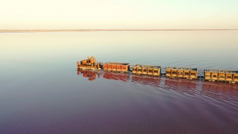 An old train travels on a railway laid in the water across a salt lake. Aerial view. Extraction of salt in Lake Bursol. Russia. September 2020