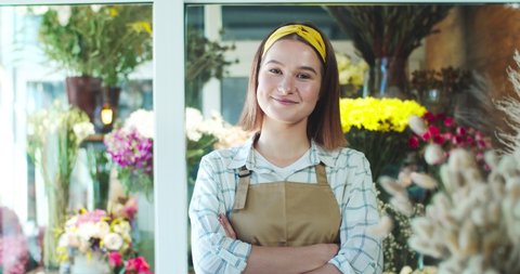 Happy Caucasian female flower shop seller wearing apron in modern store. Good-looking young woman florist crossing arms, looking at camera and smiling. Business, entreprenuership, nature concept.