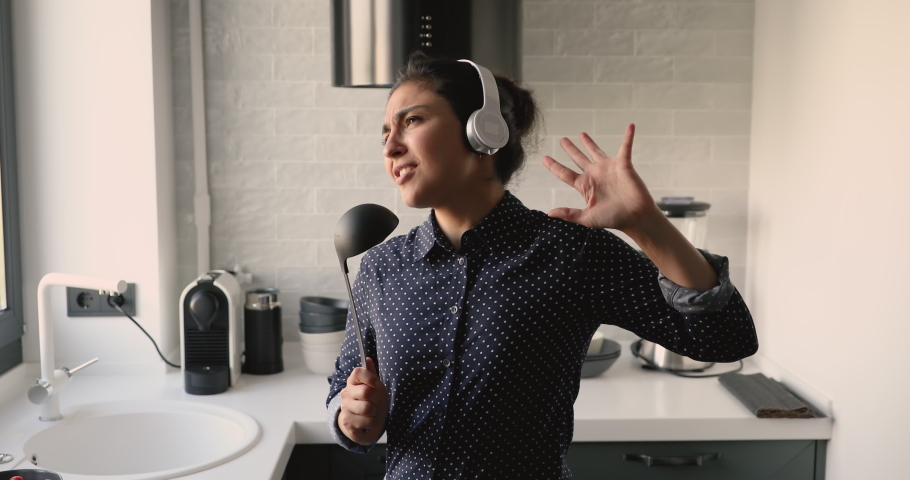 In kitchen dancing moving young indian ethnicity 30s woman, girl listens music through headphones using ladle like microphone sing favorite song enjoy life and weekend feel carefree while cook at home Royalty-Free Stock Footage #1058504104