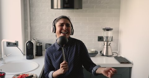 In kitchen dancing moving young indian ethnicity 30s woman, girl listens music through headphones using ladle like microphone sing favorite song enjoy life and weekend feel carefree while cook at home