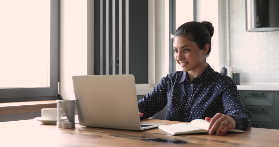 Smiling indian business lady working from home office sit at desk use laptop search information on internet writing down it in organizer agenda. Self-education, notebook personal planner usage concept | Shutterstock HD Video #1058504179