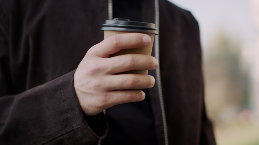 Closeup attractive man hand holding paper cup outdoors. Hipster guy hand bringing takeaway coffee to lips on city street. Handsome male person drinking tea in urban background.