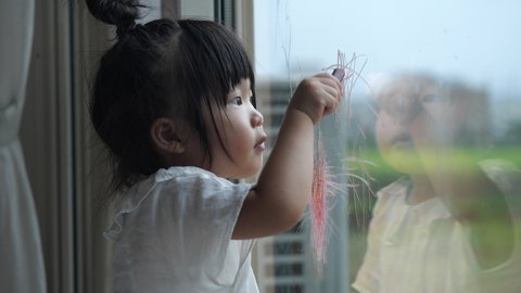 Asian preschool toddler girl in top knot hair wearing white blouse draws graffiti on the glass window with crayon. 2 years old daughter playing alone at home