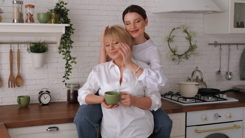 An adult daughter with an elderly mother drinks tea in the kitchen, she sits on the table and hugs her mother.