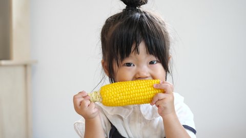 Asian preschool toddler girl in cute top knot hair eats fresh boiled corn and becomes smile. 2 years old Japanese child. Safe and healthy food. Slow motion