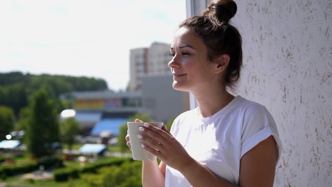 Pretty young woman on balcony drinks cup of coffee or tea watching a beautiful urban view and enjoy relax breathing fresh freezing air of summer. slow motion