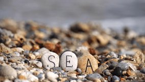 Word Sea is written on stones close-up, against background of sea stones, sea and waves. Text on beach. Letters are written on stones of sea. Relax, spa and summer vacation. Slow motion video.