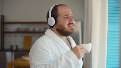 Happy man in bathrobe with cup of coffee listening to music in headphones standing near window. Funny fat guy enjoying fresh coffee and music in headphones singing and looking out of window in morning