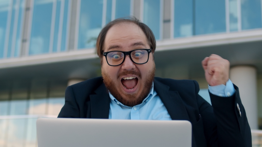 Close up excited chubby businessman doing winner gesture working on laptop outside office building. Happy overweight entrepreneur in glasses raising fist celebrating success looking at laptop screen | Shutterstock HD Video #1058519194