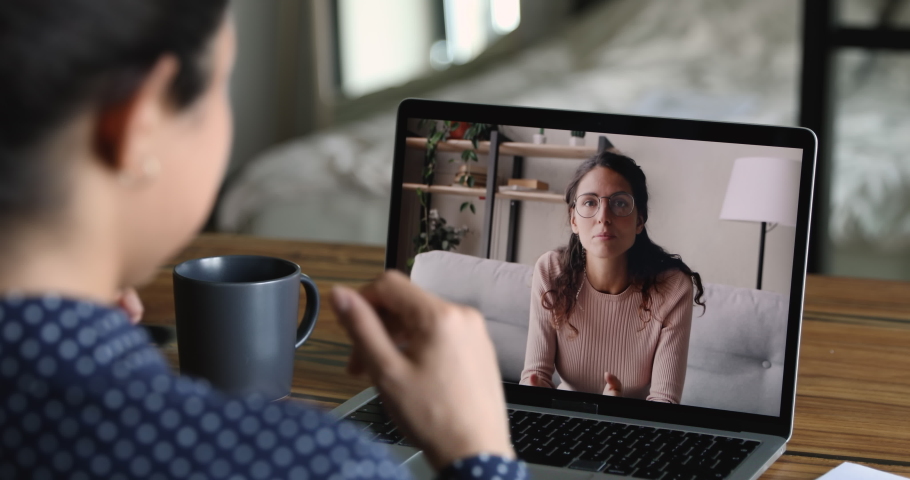 Two women colleagues discuss common project by video call application sit at desk working distantly use modern tech. View over indian female shoulder. Video conference easy comfort communication Royalty-Free Stock Footage #1058519344