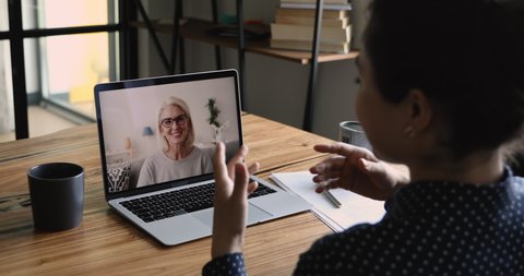 Pleasant warm distant conversation by video call laptop screen view over indian girl shoulder. Older and younger women wave hand greet each other talk. Videoconference application concept