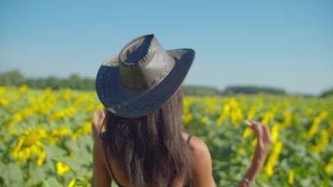 Rear view of cute carefree young african woman farmer in cowboy hat walking through blossoming sunflower field. Attractive female relaxing in summer nature, enjoying leisure in farmland.