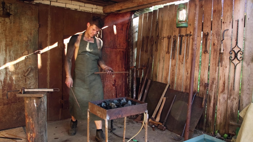A man lays metal rods in a forge. A worker in an apron does the job professionally. The skin is stained with black ash. The blacksmith is busy with his business. 4K video.