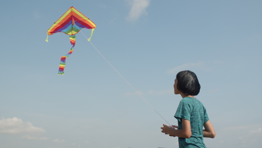 Happy Asian girl is playing rainbow kite flying on the blue sky joyful and enjoying in the summer at outdoor sunlight