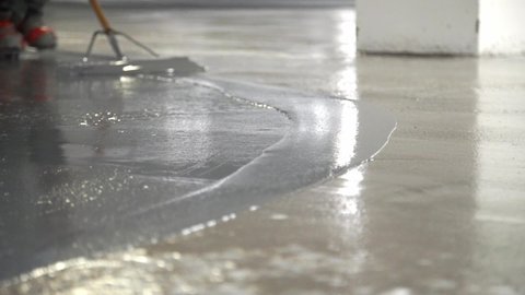 Low Angle Closeup Construction Worker Applying Gray Liquid Cement Solution Coating On Concrete Floor With Squeegee,Spike Boots Floor Leveling
