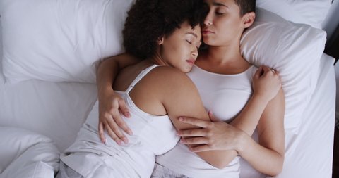 High angle side view of a mixed race female couple enjoying time at home together, lying in bed in bedroom, sleeping and embracing, slow motion