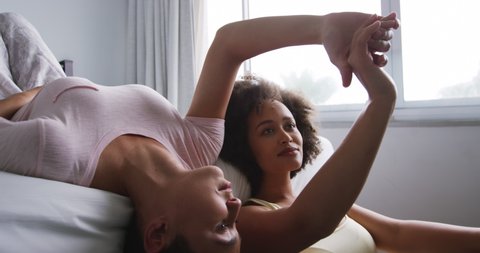 Side view of a mixed race female couple enjoying time at home together, lying on bed and sitting on a cushion on the floor in bedroom, talking holding hands, slow motion Video Stok