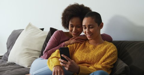 Front view of a mixed race female couple enjoying time at home together, sitting on a sofa in sitting room, taking selfies on smartphone, smiling