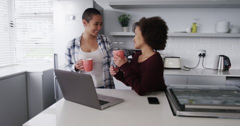 Front view of a mixed race female couple enjoying time at home together, drinking coffee and using a laptop computer in kitchen, smiling