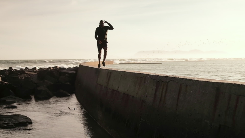 African American man wearing sports clothes, enjoying exercising at the beach, jogging on a sea wall, backlit, in slow motion