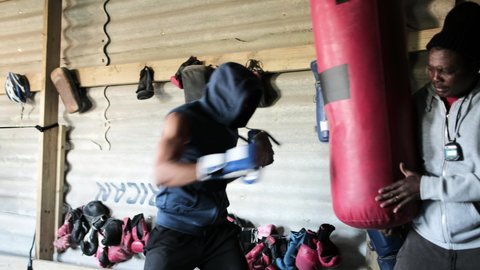 African American male boxer wearing boxing gloves training at a township boxing gym throwing punches at a punchbag held still by his African American male trainer