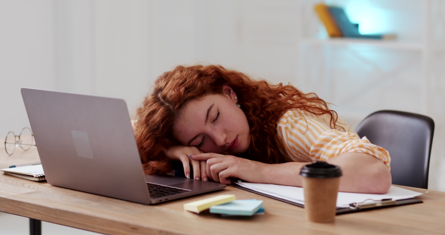 Calm relaxed young woman with red curly hair sleeping dozing peacefully on desk surface, tired of working studing on laptop, lying with head above hands.