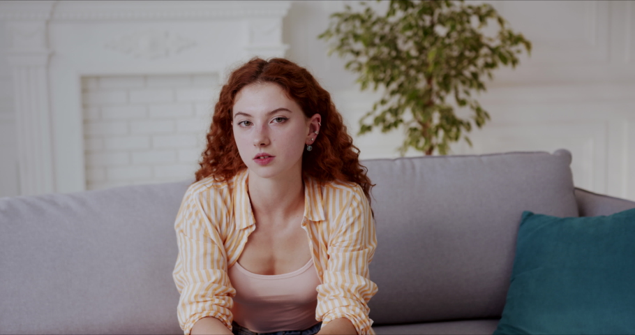 Depressed young woman with red curly hair sitting alone on sofa, thinking, having bad mood, looking upset and frustrated. Slow motion.