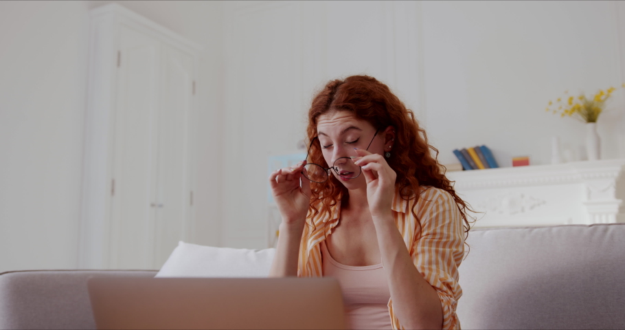 Red haired young woman tired of working or studing on laptop removes glasses feeling eye strain, rubbing dry itchy irritated eyes suffering from headache. Low angle shot.
