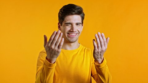 Handsome man showing - Hey you, come here. Guy asks join him, beckons with inviting hand hugs gesture. He is looking playful flirtatious, inviting to come. Yellow studio background.