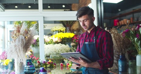 Attractive Caucasian male store owner wearing apron, working in shop. Happy young man florist using tablet to take inventory, looking at camera. Business, nature, entreprenuership concept.