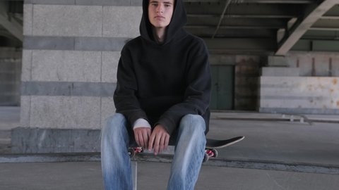 Portrait young serious not smiling caucasian skateboarder, looking at camera. Guy is sitting on skateboard in black hoodie under an iron bridge on skate Park. Slow motion video.