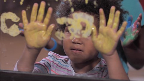 Joyful dark-skinned sisters having fun while making handprints with colored paints on home window glass. Happy curly half-breed girls enjoying palm painting during fun creative leisure