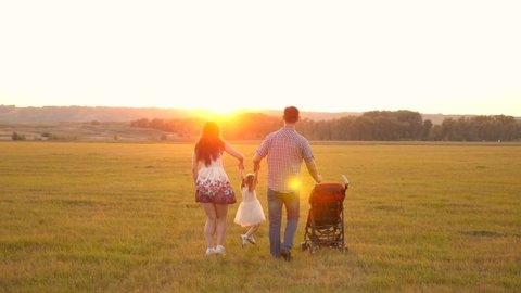 little daughter jumping, holding hands of dad and mom in park against backdrop of sun. healthy family. child plays with dad and mom on field in light of sunset. walk with a small child in nature