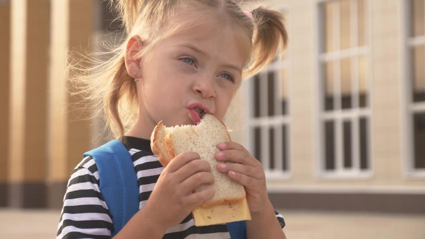 Happy kid eating a sandwich outdoors at school. Healthy school breakfast for the child. Hungry kid eating outside on the background of the school. Street fast food. Food for lunch. A delicious burger