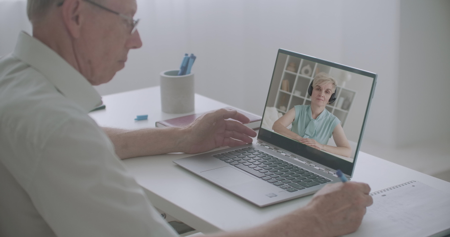 aged man is watching online training sitting at home in front of laptop, woman is lecturing, man is writing notes Royalty-Free Stock Footage #1058550931