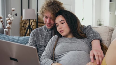 Future parenthood concept where satisfied married amorated couple of husband and pregnant woman relaxing together on the soft couch in living-room and using computer