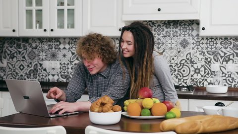 Charming smiling happy pregnant 25-aged woman coming to her busy likable curly husband which working on computer in well-equipped cuisine and he kissing her in cheek