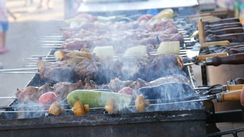 Man turn long row of vegetables and pieces of BBQ meat on skewers, ready to grill, smoke from the grill. Street food festival.