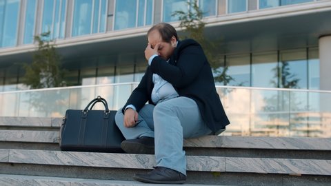 Stressed overweight businessman in formal wear sitting on street stairs worried about problem at work. Depressed fired office worker sitting on staircase outside modern business center