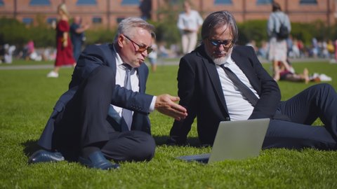 Aged business partners watching presentation and discussing project sitting on green lawn in city park. Mature executives using laptop working together in summer park