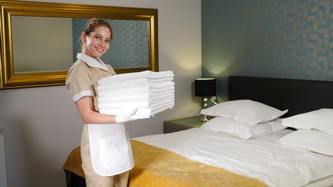 Cheerful female housekeeper holding stack of clean towels, laundry service. Hotel room preparation. Maid tidying up in hotel suite and bringing few clean fresh towels for residents. Room service. 4 k
