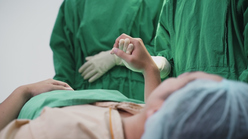Asian patient on operating theater waiting for operation by group of surgeon doctor and medic team in operation room at hospital. Doctors give encourage to inpatient by holding hand and touch shoulder | Shutterstock HD Video #1058565505