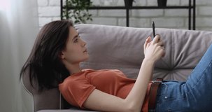 Relaxed girl lying down on couch with cellphone enjoy calm free time in living room using wireless gadget, download learn news cool app feels carefree, generation and modern technology overuse concept