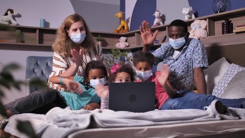 Cheerful mixed race family wearing protective masks using laptop on bed while communicating online via internet. Masked parents and girls waving hands while greeting relatives during remote connection