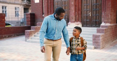 Handsome African American man walking with cute schoolboy near school and talking. Teen junior student with backpack telling something his dad. Father listening to son. Pupil concept