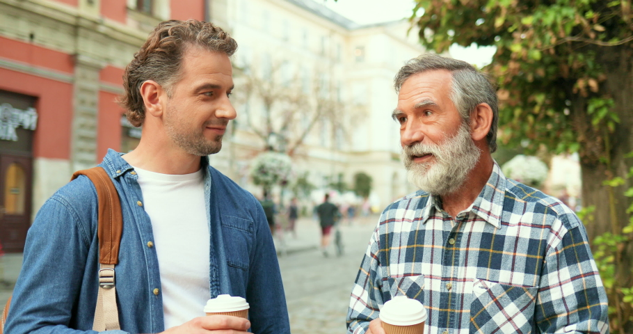 Caucasian handsome young adult man talking with old father while walking in city street and drinking coffee to-go. Two me of different ages and generations having nice conversation outside in town.