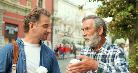 Caucasian handsome young adult man talking with old father while walking in city street and drinking coffee to-go. Two me of different ages and generations having nice conversation outside in town.