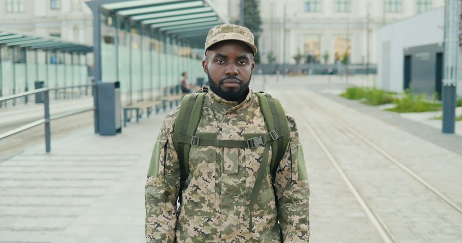 Portrait of serious African American handsome young male soldier in cap with backpack standing at street. Man militarian looking at camera at train station. Bus stop. Military uniform. Zooming in. Royalty-Free Stock Footage #1058573830