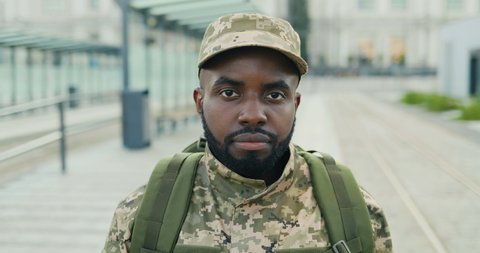 Portrait of serious African American handsome young male soldier in cap with backpack standing at street. Man militarian looking at camera at train station. Bus stop. Military uniform. Zooming in.