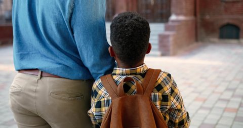Rear of handsome African American man walking with cute schoolboy near school and chatting outdoors. Close up portrait of teen male pupil with backpack walks with his dad. Family concept